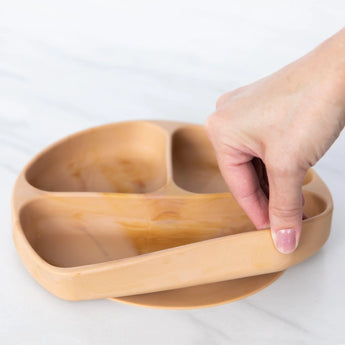 A hand holds a Bumkins Silicone Grip Dish: Wood Grain, demonstrating the suction cup bottom on a marble surface.