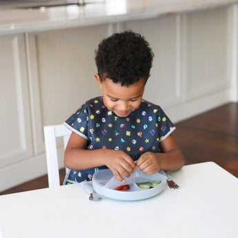 A toddler wearing a black patterned bib opens a Bumkins Silicone Grip Dish with Lid (3 Section): Gray with snacks at a white table.