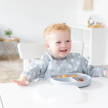 A child in a gray sleeved bib sits happily in a bright room at a white table with a Bumkins Silicone Grip Dish with Lid (3 Section): Gray.