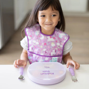 A child in a purple bib smiles at a white table with a Bumkins Silicone Grip Dish with Lid (3 Section): Lavender and matching utensils.