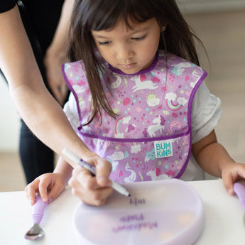 An adult writes on the lid of a Bumkins Silicone Grip Dish with Lid (3 Section): Lavender as a child in a purple bib sits nearby.