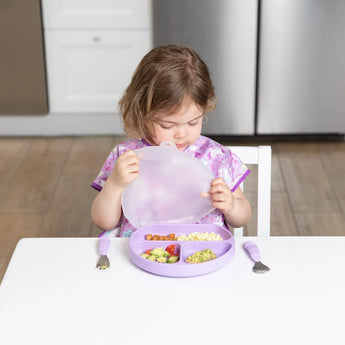 A child in a purple bib holds the lid of a Bumkins Silicone Grip Dish with Lid (3 Section): Lavender at a white table, near a fork & spoon.