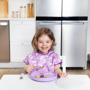 A smiling toddler in a purple bib sits at a white table with a Bumkins Silicone Grip Dish with Lid (3 Section): Lavender in a kitchen.