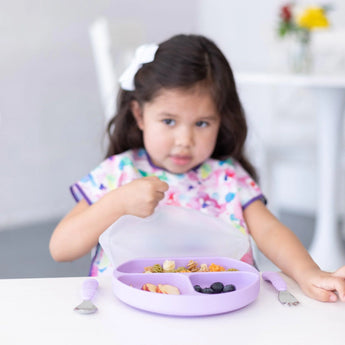A girl in a colorful bib lifts the lid of Bumkins Silicone Grip Dish with Lid (3 Section): Lavender with snacks at a white table.