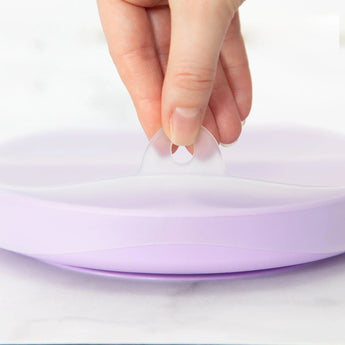 Close-up of a hand lifting the tab on the lid of Bumkins Silicone Grip Dish with Lid (3 Section): Lavender on a marble surface.