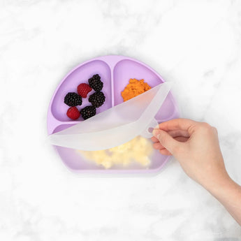 A hand lifts the lid of Bumkins Silicone Grip Dish with Lid (3 Section): Lavender, with berries, orange mash, and eggs on a marble surface.