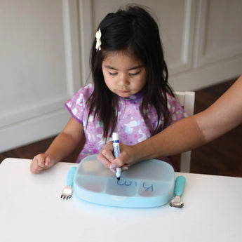 An adult hand writes on the lid of Bumkins Silicone Grip Dish with Lid (5 Section): Blue at a white table while a child sits nearby.
