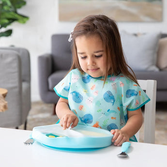 A young child in a blue bib lifts a lid from a Bumkins Silicone Grip Dish with Lid (5 Section): Blue at a white table in a cozy living room.