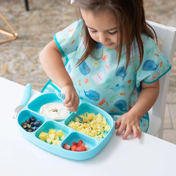 A child enjoys breakfast from a Bumkins Silicone Grip Dish with Lid (5 Section): Blue with a spoon at a white table while wearing a blue bib.
