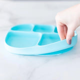 A hand holds a Bumkins Silicone Grip Dish with Lid (5 Section): Blue, demonstrating the suction cup bottom on a marble surface.