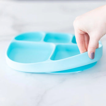 A hand holds a Bumkins Silicone Grip Dish with Lid (5 Section): Blue, demonstrating the suction cup bottom on a marble surface.