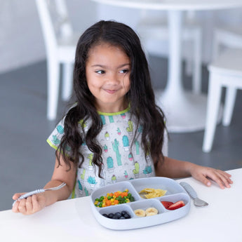 A young girl with long hair at a white table, enjoying fruits and veggies from a Bumkins Silicone Grip Dish with Lid (5 Section): Gray.