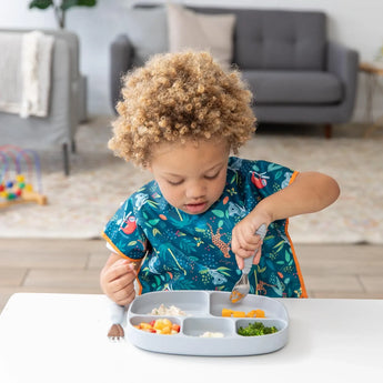 A child enjoys a meal from Bumkins Silicone Grip Dish with Lid (5 Section): Gray with a spoon, wearing a colorful bib in a cozy living room.
