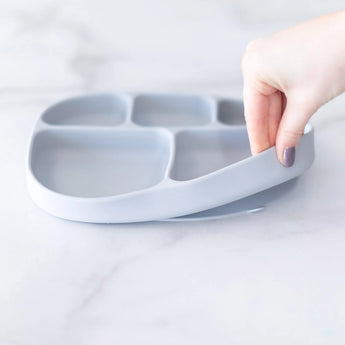 A hand holds a Bumkins Silicone Grip Dish with Lid (5 Section): Gray, demonstrating the suction cup bottom on a marble surface.