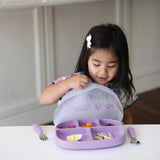 A child with long hair sits at a table, excitedly opening the lid of a Bumkins Silicone Grip Dish with Lid (5 Section): Lavender.