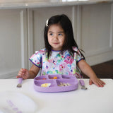 A young girl in a colorful bib sits at a white table with a Bumkins Silicone Grip Dish with Lid (5 Section): Lavender and matching utensils.