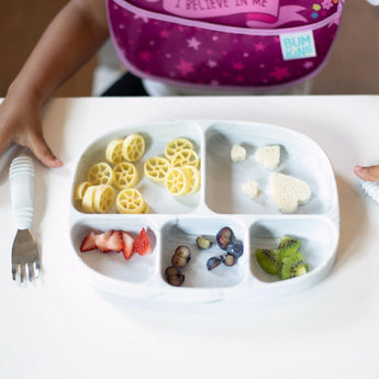 Close-up of a child at a white table with Bumkins Silicone Grip Dish with Lid (5 Section): Marble holding pasta wheels, fruit, and bread.