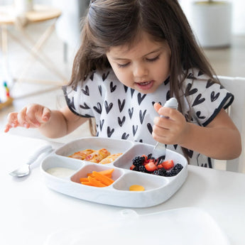 A young girl enjoys snacks from a Bumkins Silicone Grip Dish with Lid (5 Section): Marble at a white table with matching utensils.