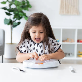 A young girl joyfully removes the lid from a Bumkins Silicone Grip Dish with Lid (5 Section): Marble at a white table in a heart-print bib.