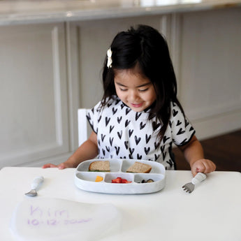 A young girl in a heart-patterned bib smiles at a meal on a Bumkins Silicone Grip Dish with Lid (5 Section): Marble at a white table.