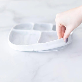 A hand holds a Bumkins Silicone Grip Dish with Lid (5 Section): Marble, demonstrating the suction cup bottom on a marble surface.