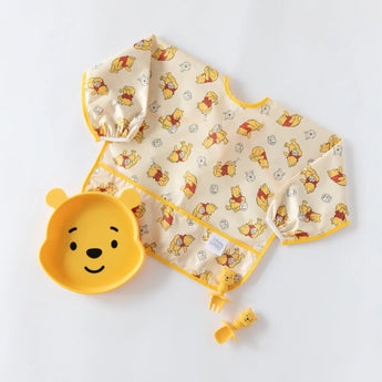A sleeved bib, Winnie the Pooh-shaped dish, and utensils from Bumkins Disney Little Ones Gift Bundle, Winnie The Pooh on a white background.