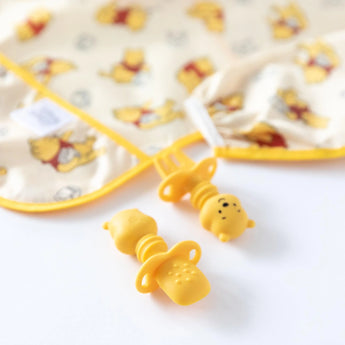 Close-up of a yellow sleeved bib and silicone utensils from Bumkins Disney Little Ones Gift Bundle, Winnie The Pooh on a white background.