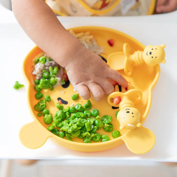 A child's hand reaches for food from a dish with utensils from Bumkins Disney Little Ones Gift Bundle, Winnie The Pooh on a high chair tray.