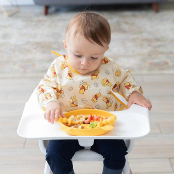 A baby in a high chair in a bib enjoys snacks from a plate with utensils, all from Bumkins Disney Little Ones Gift Bundle, Winnie The Pooh.