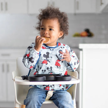 A toddler in a bib enjoys snacks from a dish while in a high chair, all from Bumkins Disney Little Ones Gift Bundle: Mickey Mouse Classic.