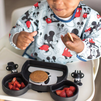 A child enjoys a pancake and berries from a plate while wearing a bib, all from Bumkins Disney Little Ones Gift Bundle: Mickey Mouse Classic.