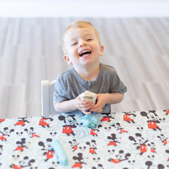 A toddler laughs, playing with clay at a table covered in a splat mat from Bumkins Disney Little Ones Gift Bundle: Mickey Mouse Classic.