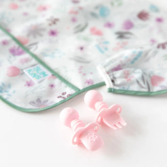 Close-up of pink silicone utensils and a floral sleeved bib from Bumkins Ultimate Gift Set - Floral & Lace on a white background.