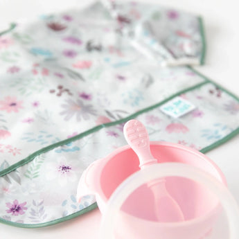 Close-up of a pink bowl, pink spoon, clear lid, and floral sleeved bib from Bumkins Ultimate Gift Set - Floral & Lace on a white background.