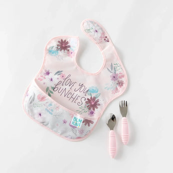 A pink floral bib and pink utensils from Bumkins Ultimate Gift Set - Floral & Lace lay on a white background.