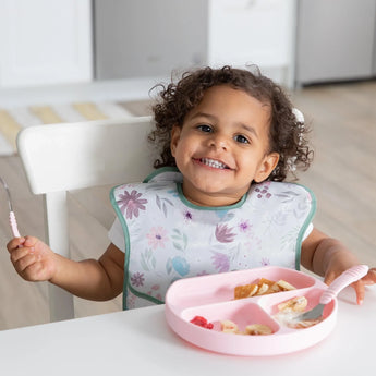 A smiling toddler wearing a floral bib eats fruit from a pink dish with utensils, all from Bumkins Ultimate Gift Set - Floral & Lace.