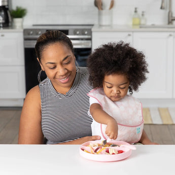 A woman smiles at a toddler wearing a pink bib, eating from a plate with utensils, all from Bumkins Ultimate Gift Set - Floral & Lace.