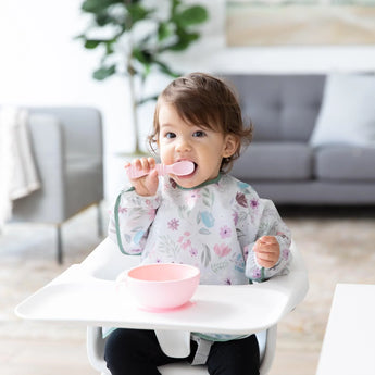 A toddler in a high chair in a floral sleeved bib eats from a pink bowl with a spoon, all from Bumkins Ultimate Gift Set - Floral & Lace.