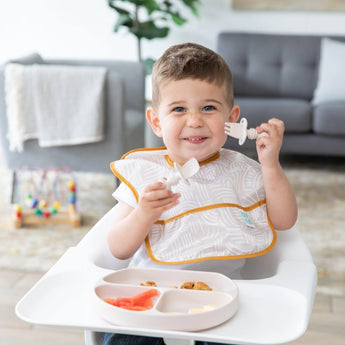 A smiling toddler in a highchair, using silicone utensils and a plate while wearing a bib from Bumkins Ultimate Gift Bundle, Desert Boho.
