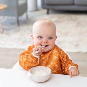 A baby at a white table wearing a sleeved bib eating from a bowl from the Bumkins Ultimate Gift Bundle, Desert Boho.