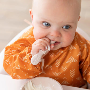 A baby in an orange bib eats from a beige bowl from Bumkins Ultimate Gift Bundle, Desert Boho while seated in a high chair.