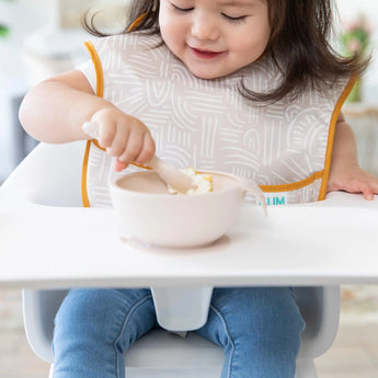 A toddler in a high chair wearing a beige bib eats from a beige bowl from Bumkins Ultimate Gift Bundle, Desert Boho.