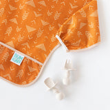 Close-up of an orange sleeved bib and beige utensils from Bumkins Ultimate Gift Bundle, Desert Boho on a white background.