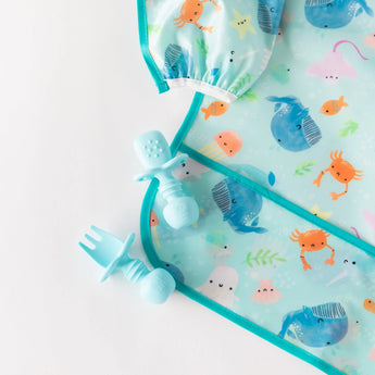 Close-up of blue utensils and a sea-themed sleeved bib from Bumkins Ultimate Gift Set - Ocean Life & Whale Tail on a white background.