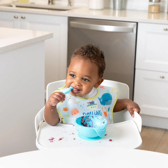 A smiling baby in an ocean-themed bib self-feeds from a blue bowl with a spoon, all from Bumkins Ultimate Gift Set - Ocean Life & Whale Tail.