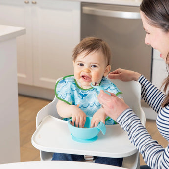 A woman helps a baby in a high chair in a sea-themed bib eat from a blue bowl, all from Bumkins Ultimate Gift Set - Ocean Life & Whale Tail.