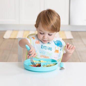 A toddler in an ocean-themed bib eats from a blue plate with a fork, all from Bumkins Ultimate Gift Set - Ocean Life & Whale Tail.