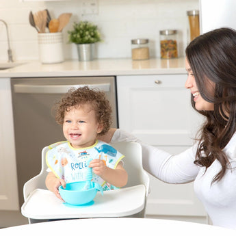 A toddler in a high chair in a bib eats from a bowl, all from Bumkins Ultimate Gift Set - Ocean Life & Whale Tail, with a woman nearby.