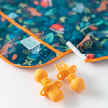 Close-up of orange silicone utensils and a jungle-themed bib from Bumkins Ultimate Gift Set - Jungle & Animal Prints on a white background.