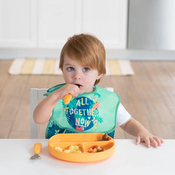 A toddler in a green bib eats from an orange dish with matching utensils, all from Bumkins Ultimate Gift Set - Jungle & Animal Prints.